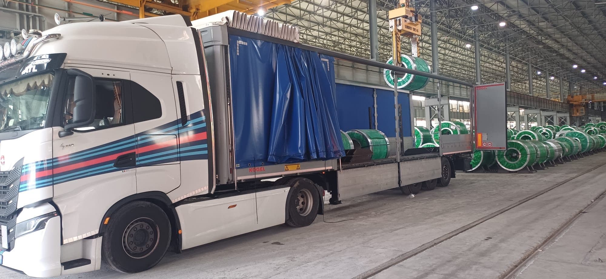 Trattore Iveco con semirimorchio telonato Kögel aperto su un lato dentro un'acciaieria, mentre il carroponte cala un coil di acciaio nel vano di carico