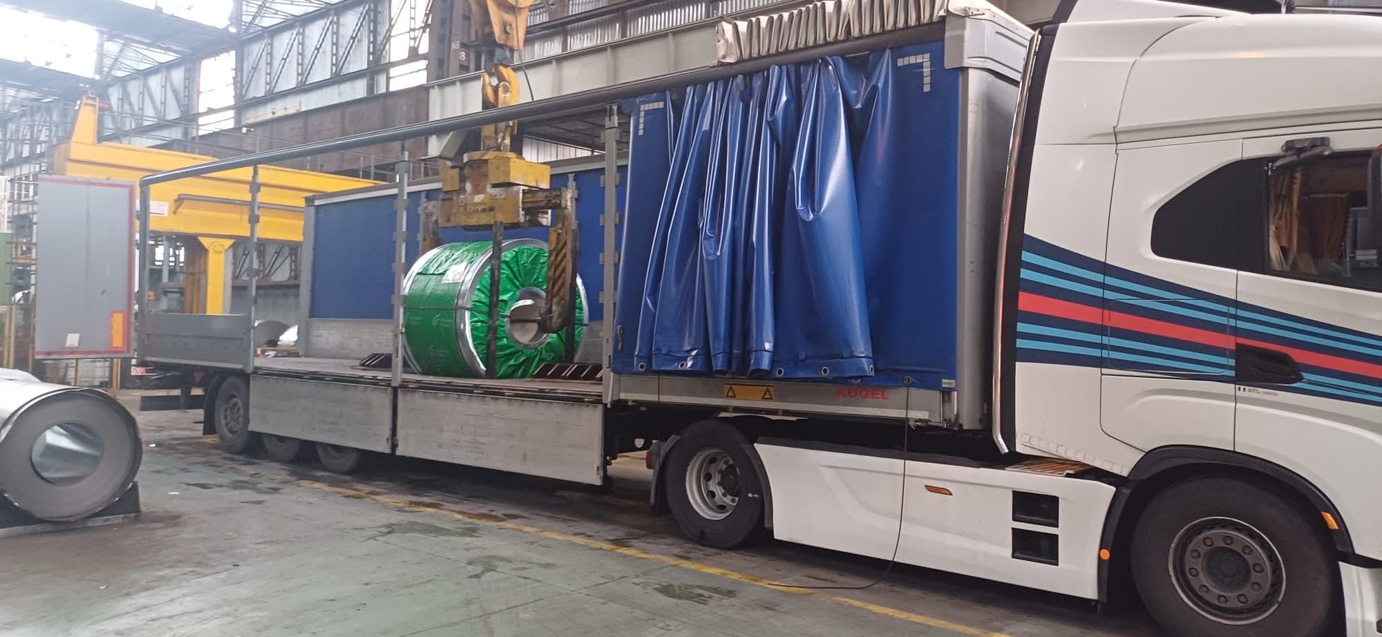Coil di acciaio imballato agganciato alla pinza del carroponte, calato dall'alto nel semirimorchio telonato con la tenda laterale aperta