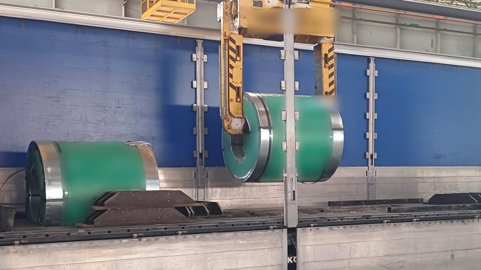 Semirimorchio telonato aperto sul fianco in un capannone: un coil è già nella culla e un secondo coil scende agganciato alla pinza del carroponte